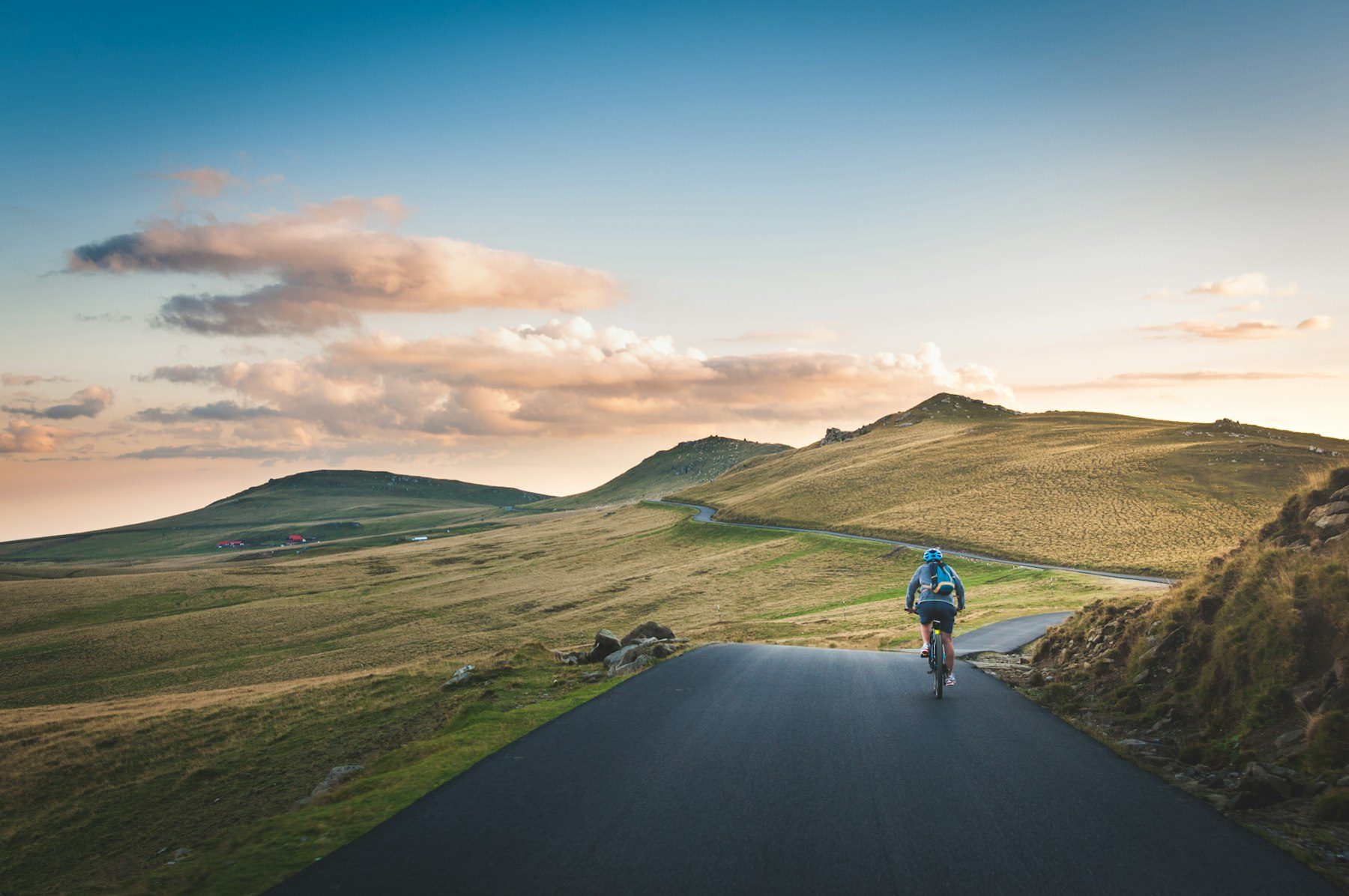 A lone cyclist rides a winding open road through golden high-country hills at sunset