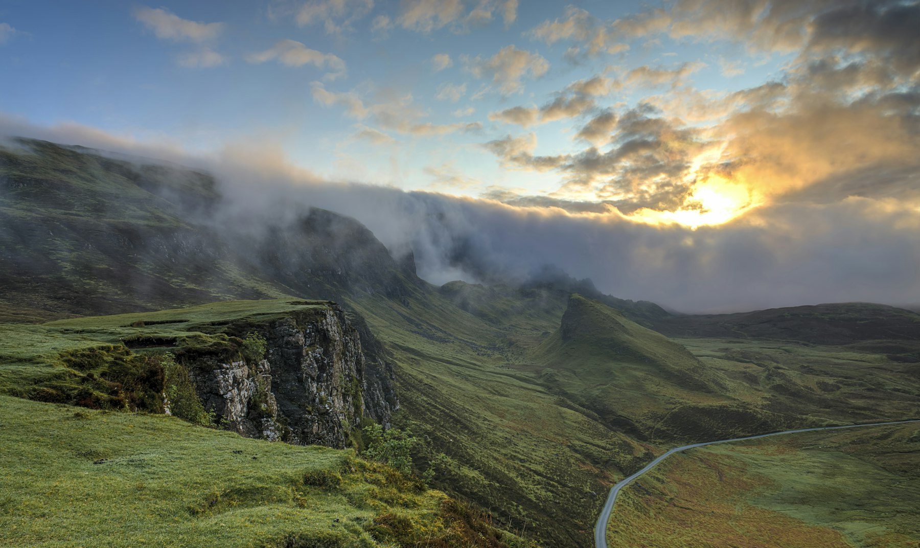 Morning sun breaks through low cloud over green highland ridges and a winding road