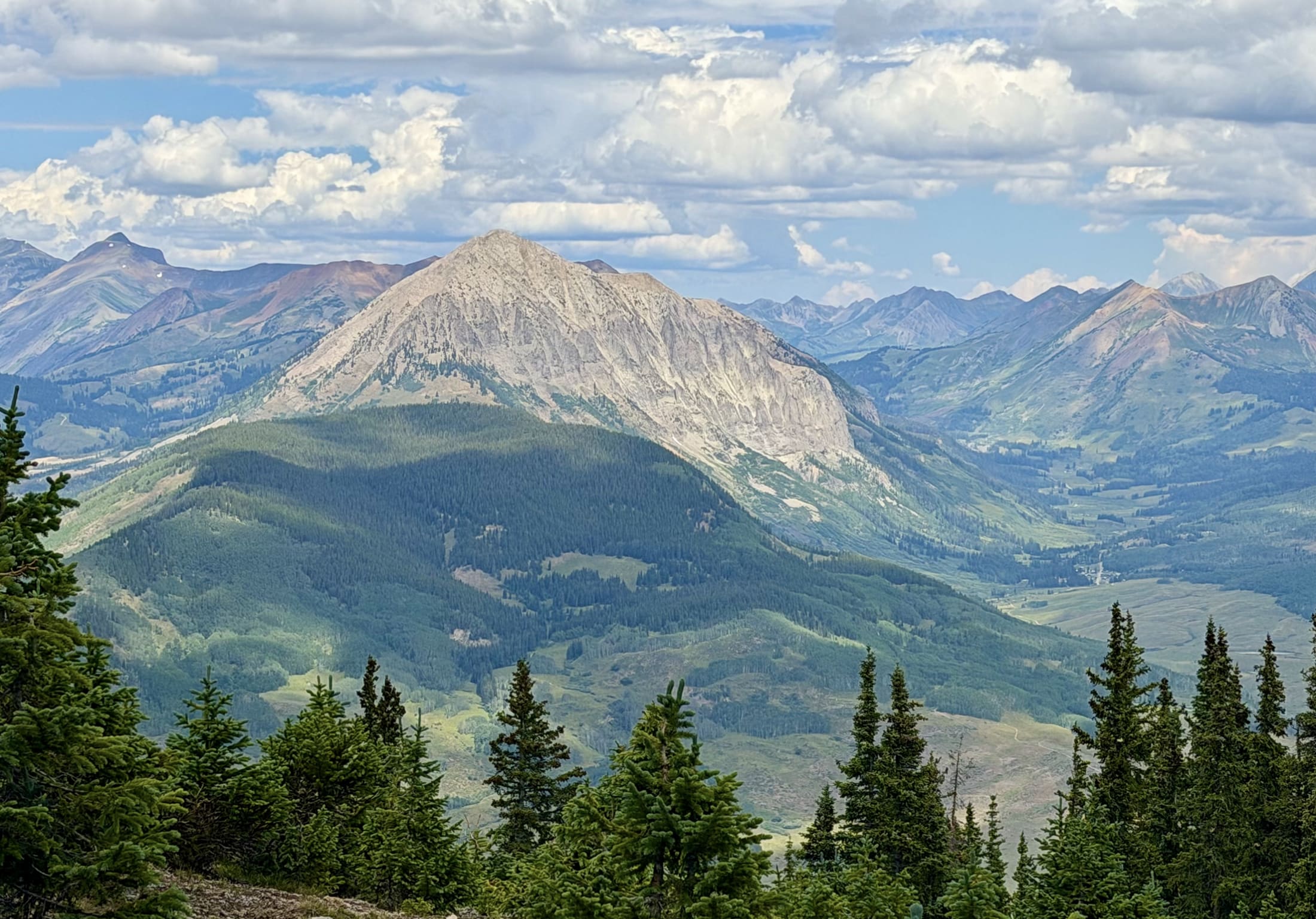 Rocky summit rising above dark evergreen forest and a green valley, under a sky of scattered clouds