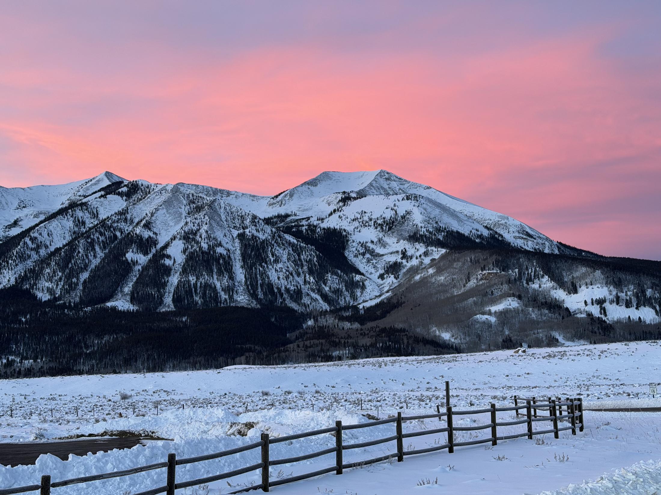 Pink sunrise sky over snow-covered peaks, with a wooden ranch fence crossing a snowy meadow in the foreground